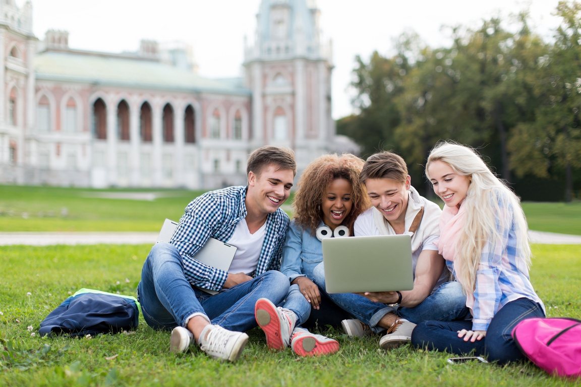 Four students sitting on a campus lawn, sharing a laptop